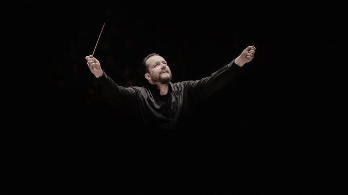 Andris Nelsons with his arms raised. He's holding a conductor's baton in his right arm, and he appears to be coming out of the black background.