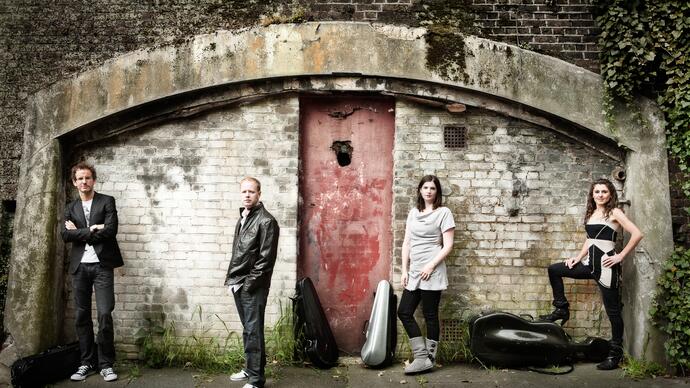 The Carducci Quartet standing in a line in front of an old, dilapidated building, their instruments in cases on the floor beside them.