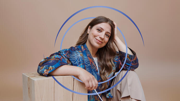 Dalia Stasevska sitting down, resting her arm on a wooden block. There are blue circular swirls around her.