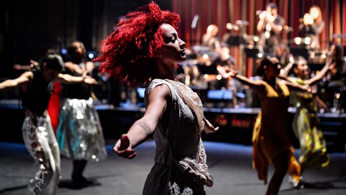 A person with red hair dances on stage with their arms outstretched. Other dancers move behind them and a jazz band plays.