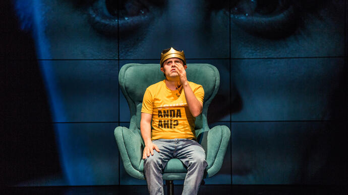 A person in a yellow shirt and blue jeans sitting on a green sofa chair with one of their hands up. They are against a background of screens with a face on it 