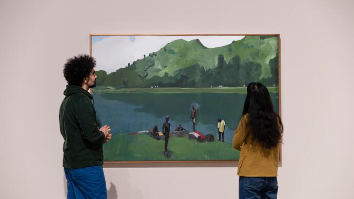 A man and a woman stand in front of a large painting depicting people lounging on the grass next to a large body of water.
