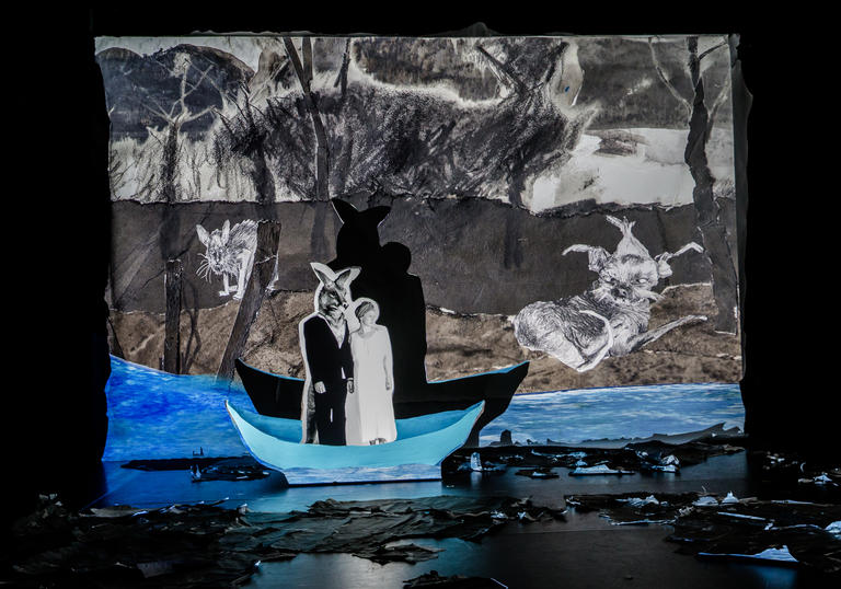 A figure that is half-man, half-kangaroo is standing in a boat with Fleur Elise Nobel's unnamed character. They look like figures in a pop-up book, against a paper painted backdrop and vivid blue colour in the water. Their shadows are prevalent against the paper backdrop.