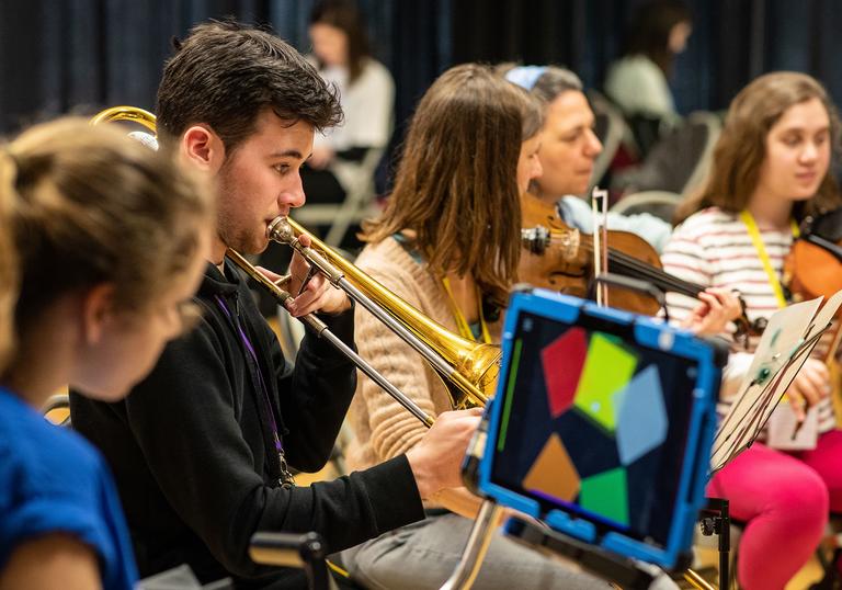 A group of young people in a orchestra, one boy is playing the trumpet and two girls in the background are playing the violin.