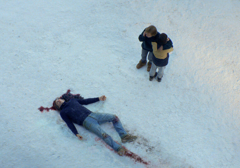 An aerial shot of a man lying in the snow, with blood on his head. Someone crouches nearby looking at him.