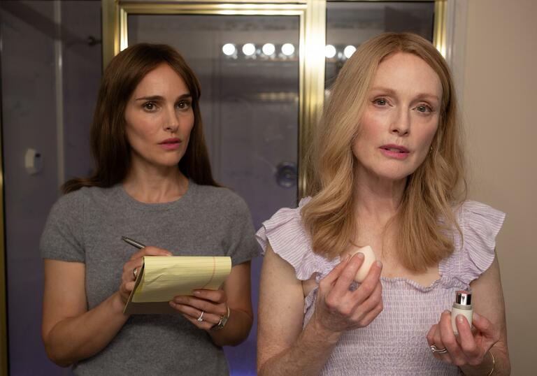 Two women look in a mirror; one puts on makeup, the other writes in a notepad.