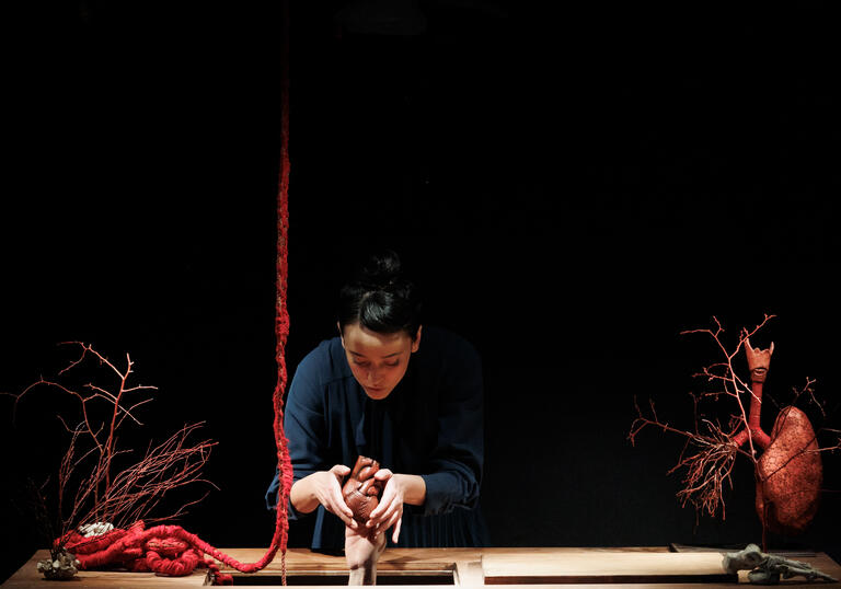 A person stands at a wooden work bench and moulds a small human body out of clay. A large red rope hangs from the ceiling like intestines.