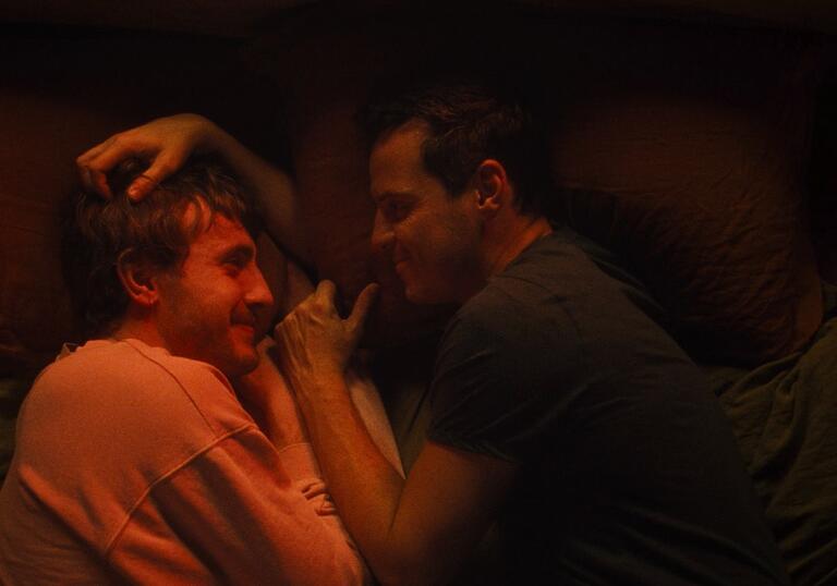 Two men lie in bed together, draped in orange light. They look lovingly at each other.