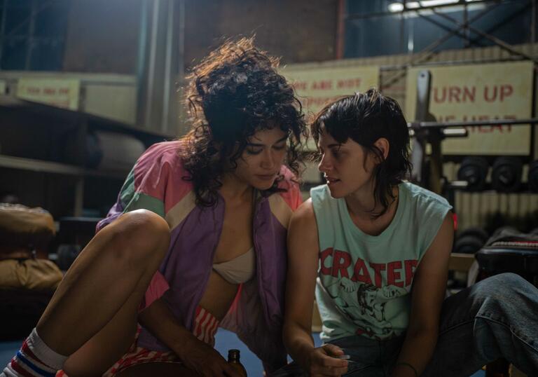 Two women in gym clothes sit on the floor of a boxing ring, holding hands.