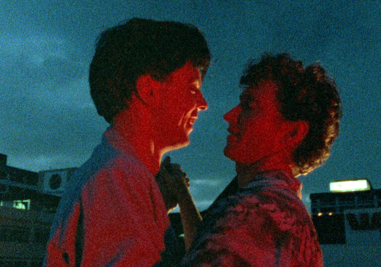 Two young men embrace each other in a dark city skyline.