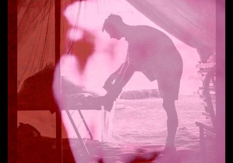 A pink and red image of a man tying his shoe lace in what looks like an army tent in a field.