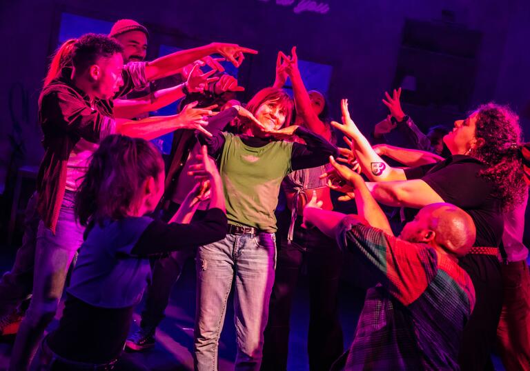 A group of performers gather around one performer and raise their hands towards her.