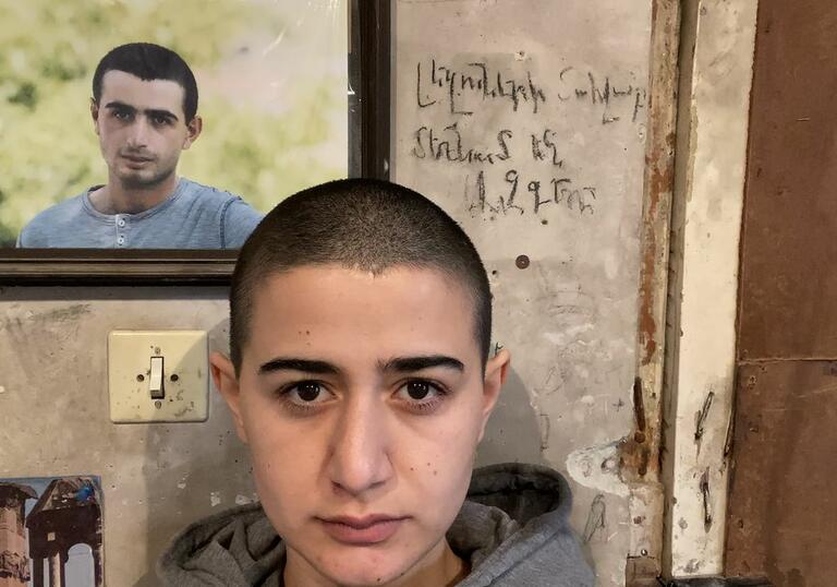 A young boy stands against a wall, that holds a photo of an older boy. 