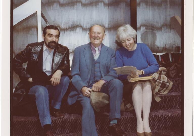 Two men and a woman who is reading a book and laughing sit on a wall together in this polaroid image.