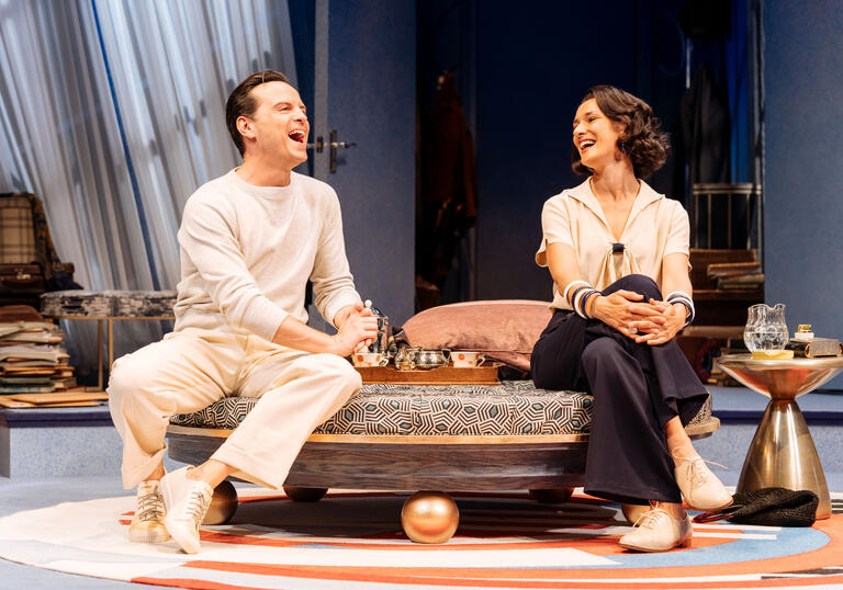 A man and a woman sit on a divan on stage laughing with each other.