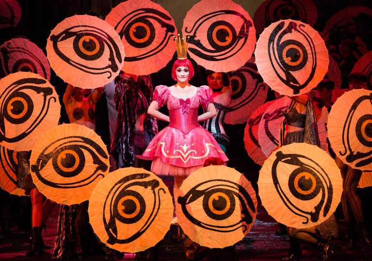 An actor stands in the middle of the stage in a pink dress, surrounded by large eyes.