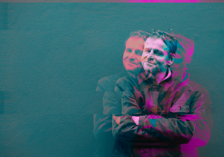 Writer William Adamsdale poses with his arms crossed against a blue background. There is a pink effect around him that blurs.