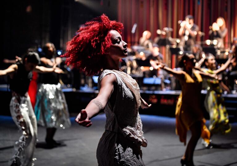 A person with red hair dances on stage with their arms outstretched. Other dancers move behind them and a jazz band plays.