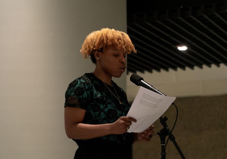 A Barbican Young Poet holds a piece of paper and reads into a microphone.