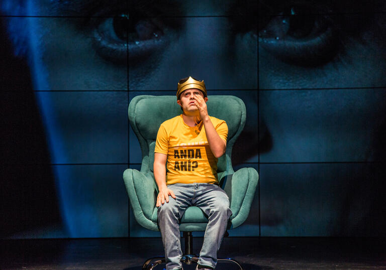 A person in a yellow shirt and blue jeans sitting on a green sofa chair with one of their hands up. They are against a background of screens with a face on it