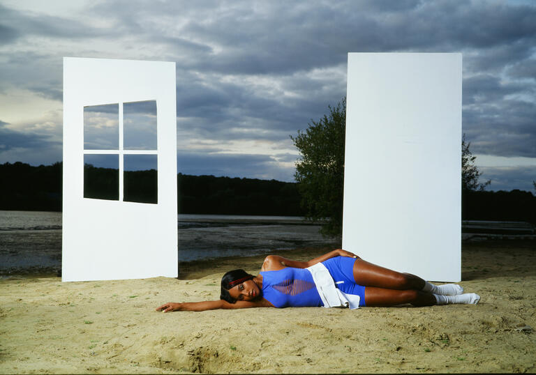 A person lies down on the beach in a blue dress. Two white walls stand behind them, one with a window. The sea can be seen in the background along with the sky.