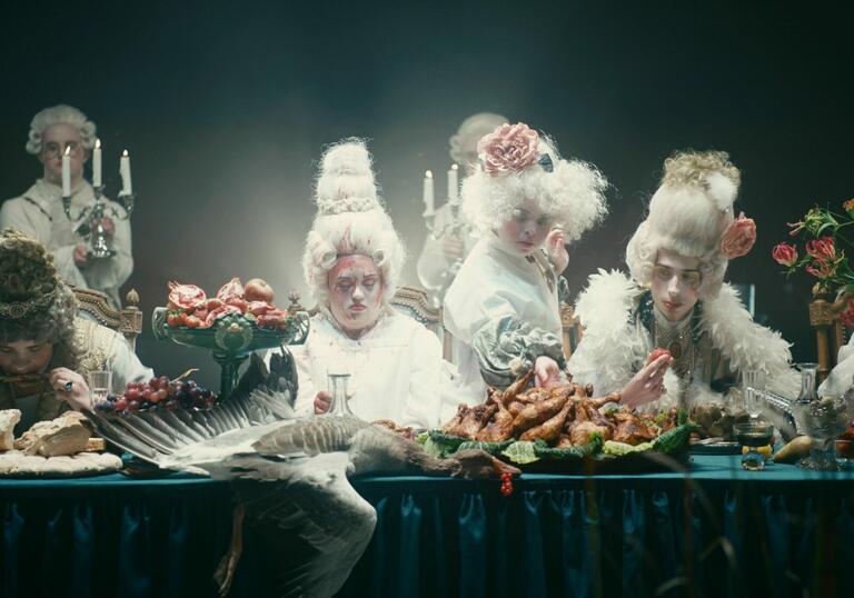 A group of actors in wigs sit at a table covered in food.