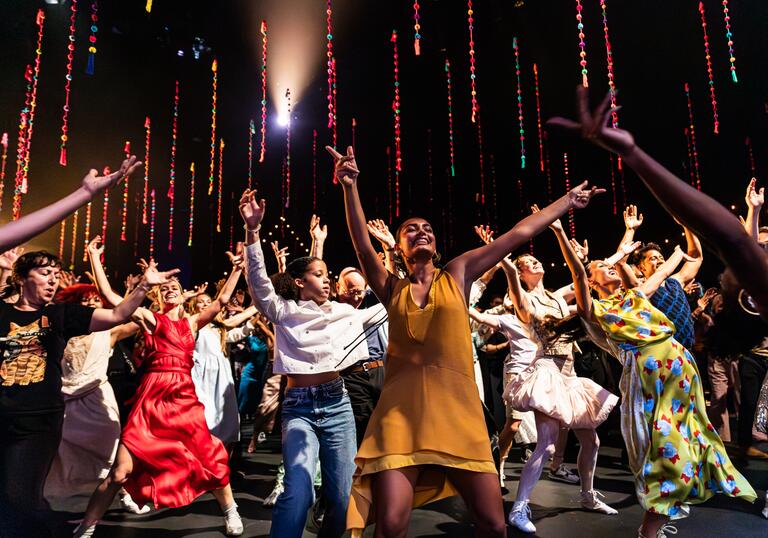 A group of people wearing colourful clothing dance enthusiastically under beads that hang from the ceiling