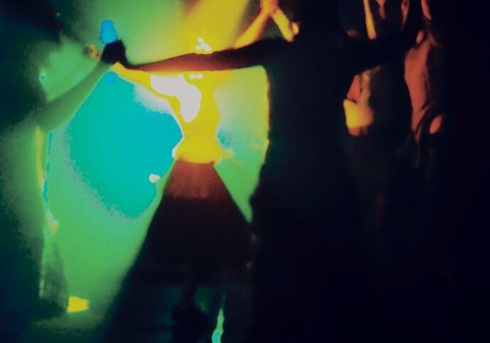 Figures are dancing in a dark room lit by yellow, green and blue lights.