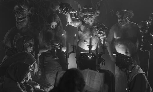 A satanic group gather in dark candlelight.