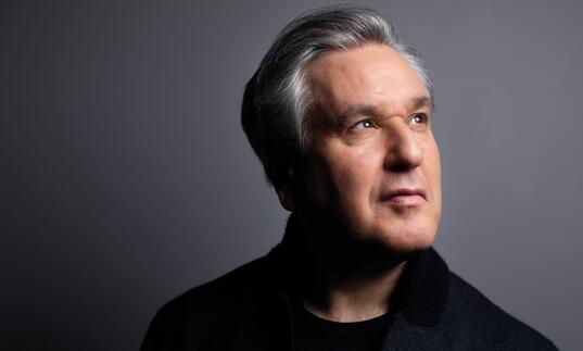 A portrait photo of Sir Antonio Pappano, against a grey background, looking away from the camera into the distance.