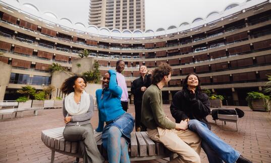 A group of six young people sat and standing, laughing in the Sculpture Court at the Barbican