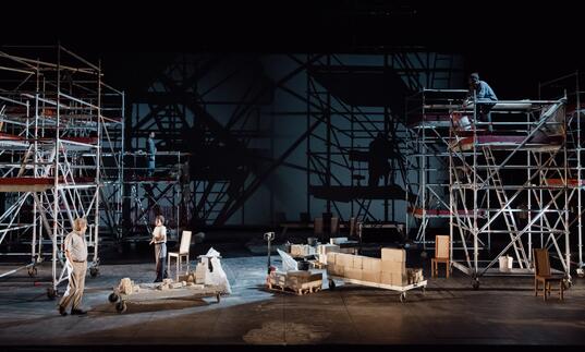 Two people stand on stage with a minimal set of furniture while tall scaffold towers line the edge of the stage.