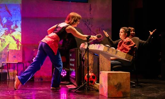 Flo reaches over the tech desk to Rachel with a microphone.