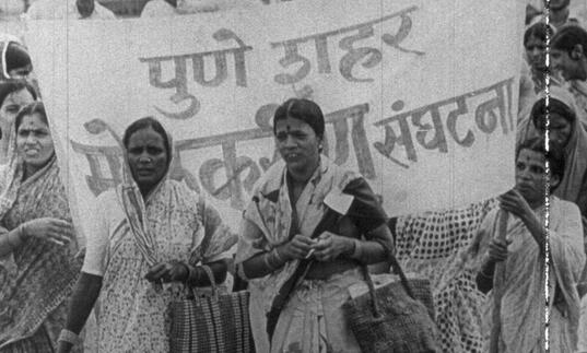 Women stand infront of a protest banner marching down the street.
